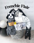 A baby gift basket titled 'Frenchie Flair' containing a black and white onesie, a matching blanket, a plush toy resembling a French Bulldog, wash cloths, socks, a hooded towel, and a fabric basket.