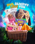 A wicker basket gift set with owl-themed baby clothing and accessories, including a furry vest, long-sleeve top, pants, socks, a hat, hooded towel, and a children's book, all arranged with a nature and owl aesthetic.