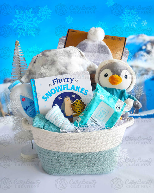 A winter-themed gift basket containing a plushy penguin toy, a book titled 'Flurry of Snowflakes', a grey onesie, a fleecy soft plaid blanket, a matching pompom hat, baby powder, wet wipes, face cloths, and a fabric basket.