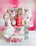 A baby gift basket with various items including a onesie, mittens, a hat, a zebra-striped blanket, a plush zebra toy, wash cloths, a milk bottle, and a food bib, all arranged in a white wooden crate.