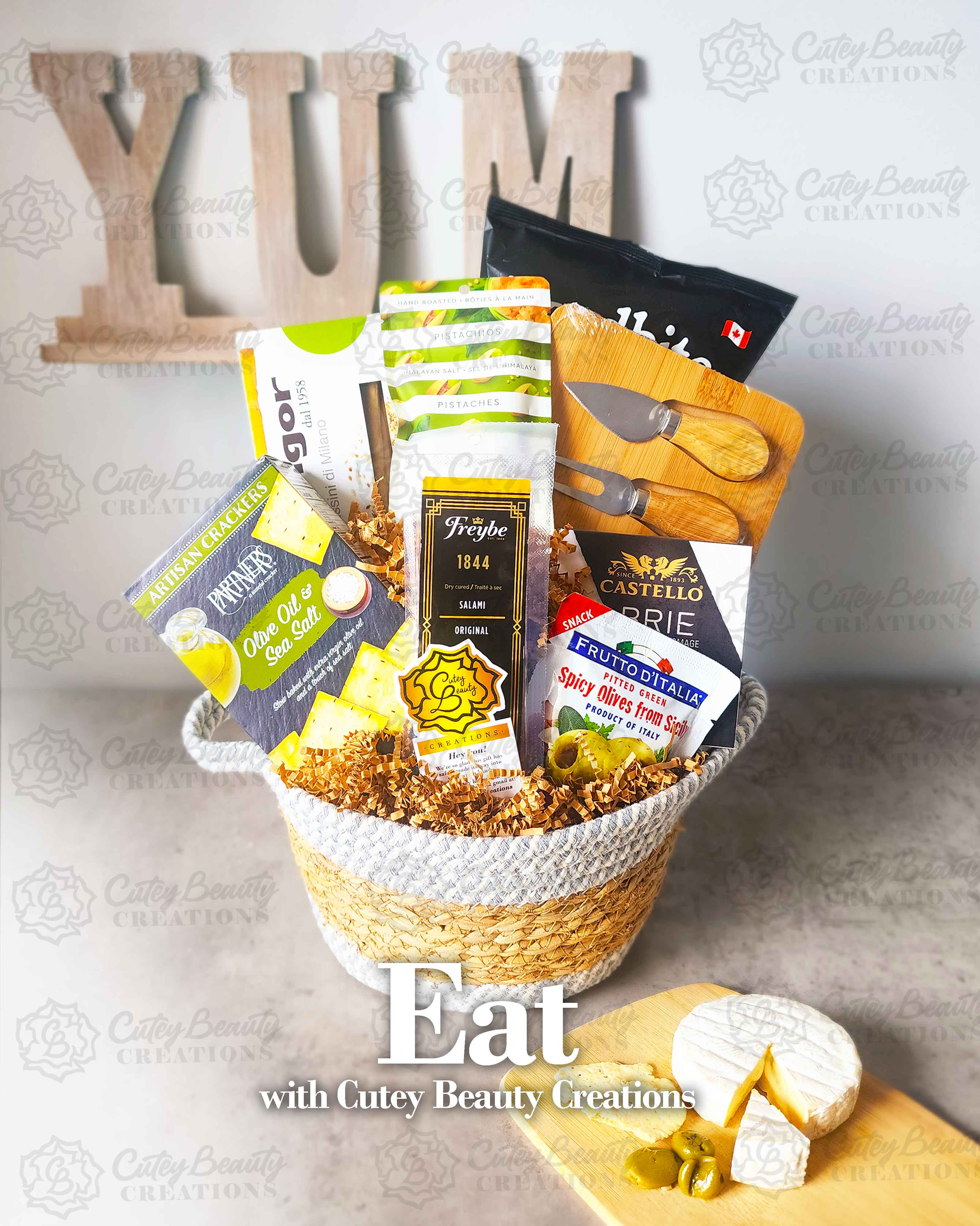 A wicker basket containing a charcuterie board set, including crackers, cheese, nuts, olives, and meats, with 'YUM' and 'EAT' signs in the background, and a piece of brie cheese on a wooden surface in the foreground.
